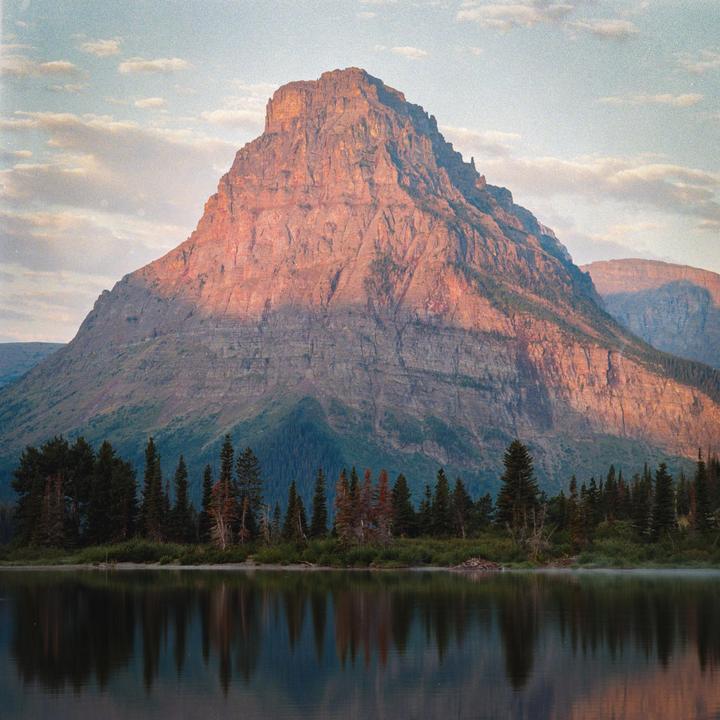 A large mountain with a sunlit peak rises above a line of evergreen trees and a calm lake reflecting the landscape.