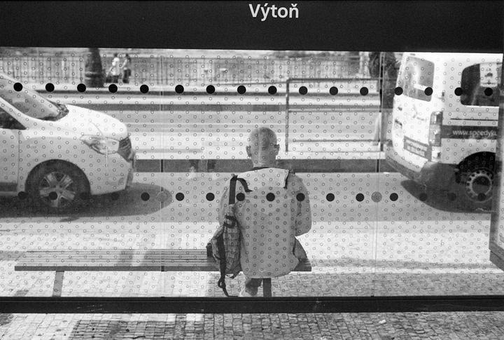 A person sits on a bench at a bus stop, seen through a patterned glass, with cars and a van passing by on the street.