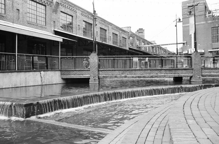 A brick building with awnings overlooks a canal with a small waterfall and a brick walkway.