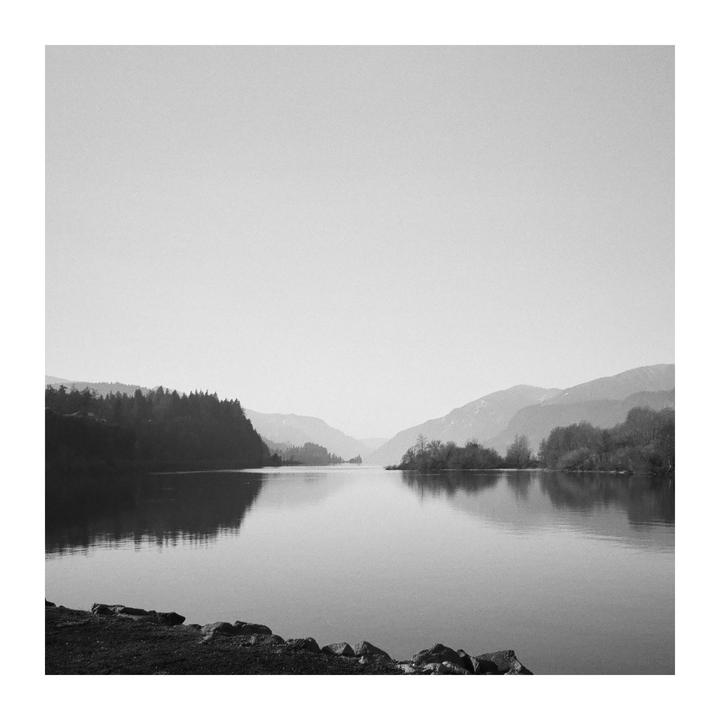 A body of water reflects the surrounding mountains and trees under a hazy sky.