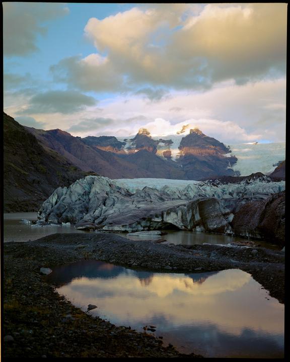 A glacier and mountains are reflected in a body of water under a cloudy sky.