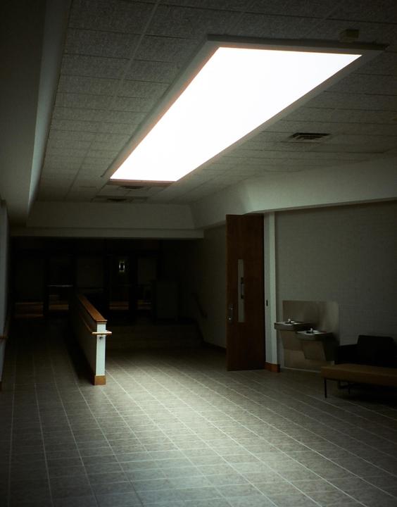 A long fluorescent light fixture illuminates a tiled hallway with a water fountain and a dark doorway.