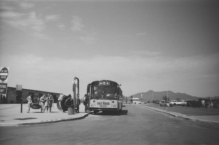 A group of people with inner tubes stand next to a bus on a sunny day with mountains in the background.
