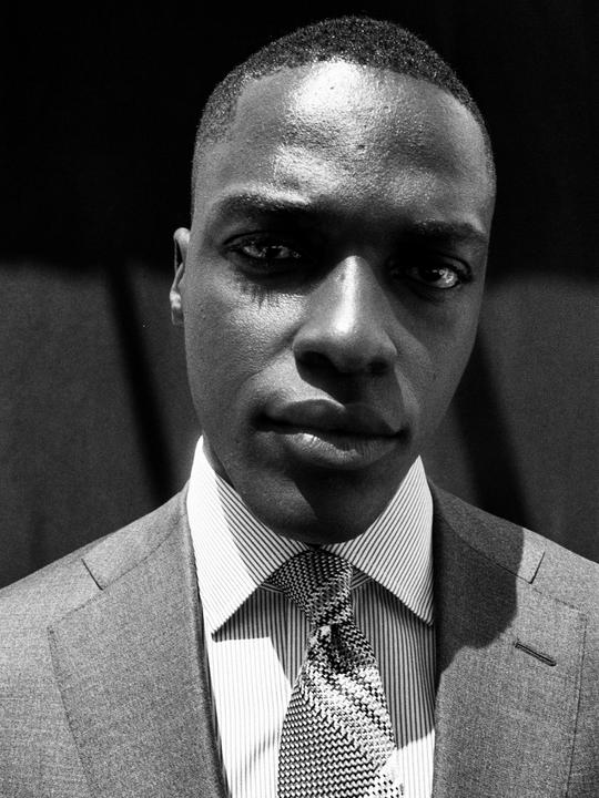 A man in a suit and striped shirt with a patterned tie looks directly at the viewer.