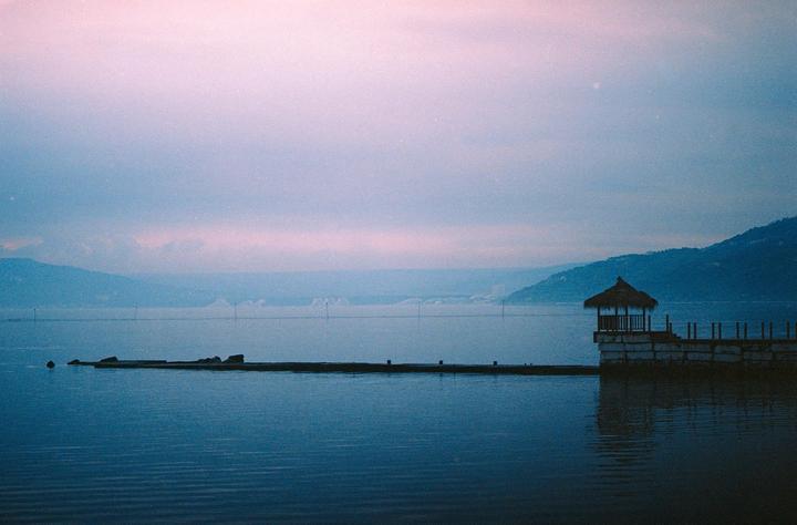 A small hut with a thatched roof sits on a pier extending into a hazy sea with mountains in the distance.