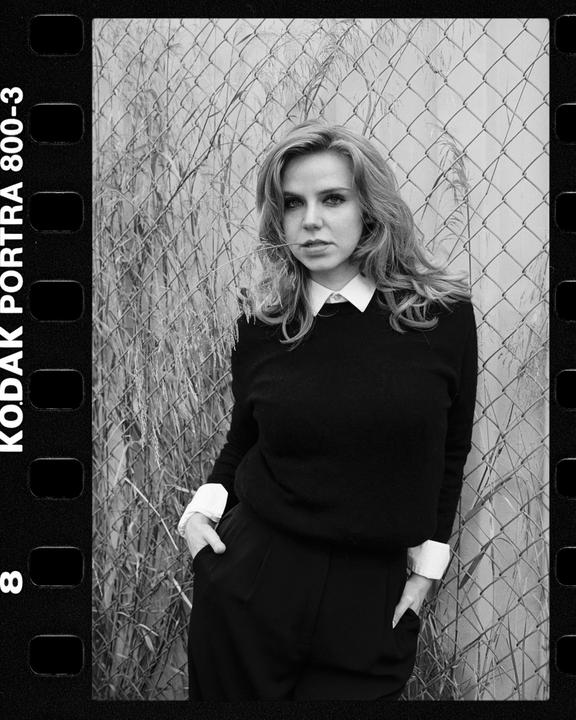 A woman with long hair and a collared shirt stands in front of a chain link fence with tall grass.