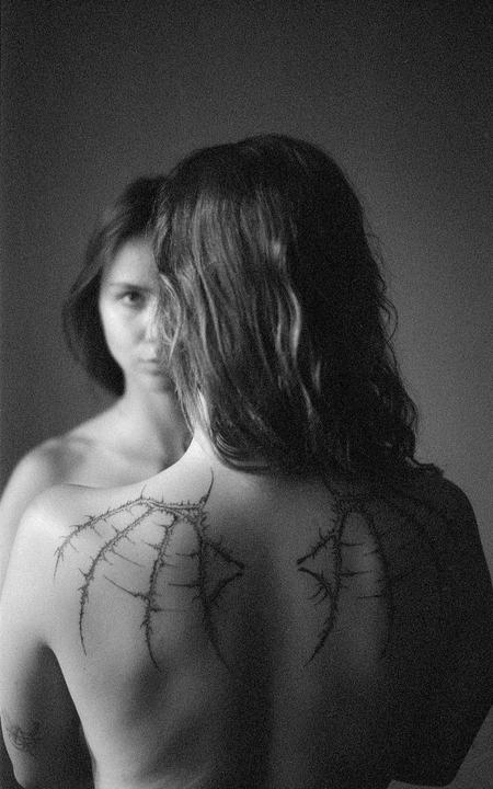 A woman with long hair and a tattoo on her back looks into a mirror, revealing her reflection looking back.