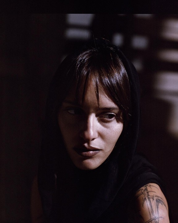 A woman with dark hair and a black hood looks to the side in a dimly lit setting.