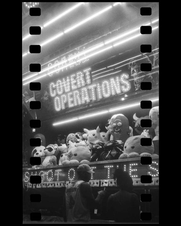 A black and white photo shows a carnival game booth with stuffed animals and two people in the foreground.