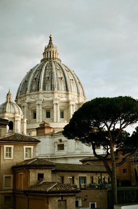 A large domed building with smaller buildings and a tree in the foreground is visible under a cloudy sky.
