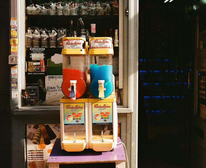 Two slushie machines with red and blue drinks sit on a table outside a shop.