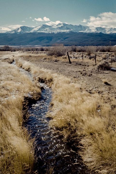 A small stream flows through a field of golden grass with snow-capped mountains in the background.