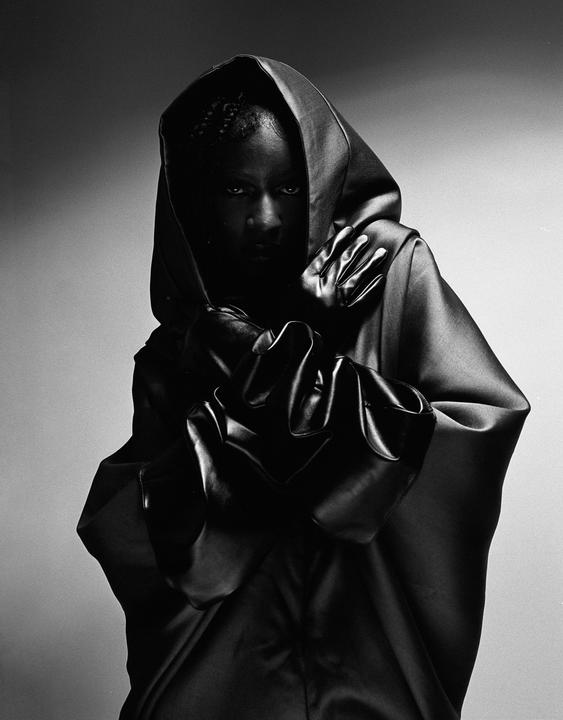 A person in a dark hooded garment and gloves looks directly at the viewer.