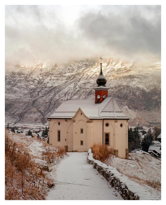 A snow-covered path leads to a white church with a red and black steeple, set against a backdrop of snowy mountains under a cloudy sky.