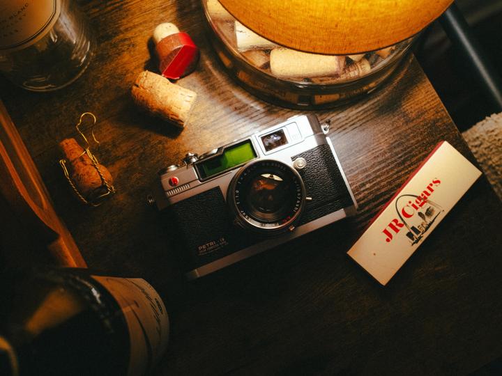 A camera, cigar box, and wine corks are arranged on a wooden surface under warm light.