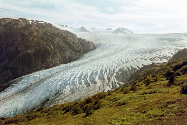 A vast glacier flows between two mountainsides, with snow-capped peaks visible in the distance under a cloudy sky.