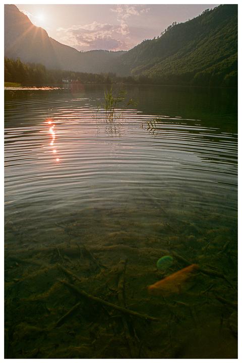 The sun sets over a lake surrounded by mountains, with the sun's reflection visible on the water's surface and submerged branches visible below.