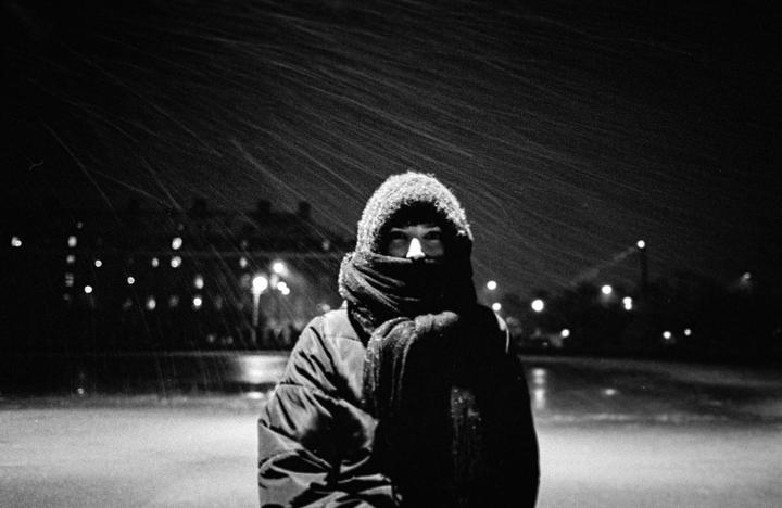 A person bundled in a hooded jacket and scarf stands in falling snow at night, with blurred lights in the background.