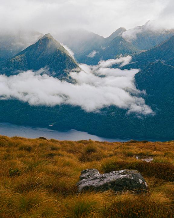 Clouds roll over a mountain range and a lake, with a field of tall grass and a rock in the foreground.