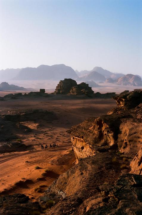 A caravan of camels and riders travels through a vast desert landscape with mountains in the distance.
