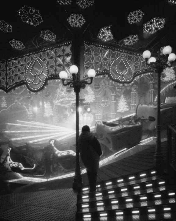 A person stands on illuminated stairs looking at a brightly lit funfair ride at night.