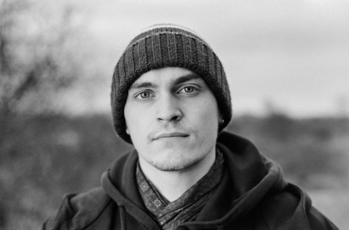 A man wearing a beanie and a hooded jacket looks directly at the viewer.