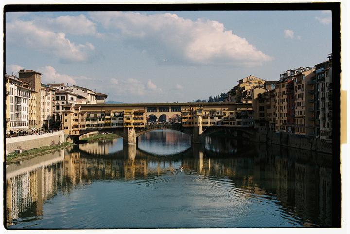 A bridge with buildings on top spans a river, with more buildings lining the riverbanks under a cloudy sky.