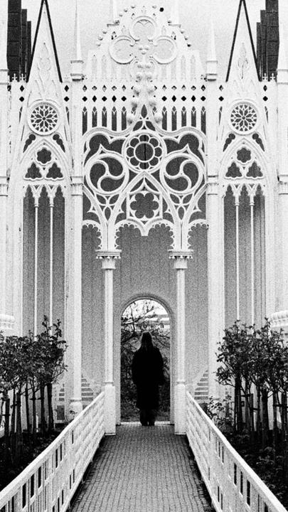 A person in a dark coat walks away from the camera through an ornate white archway.