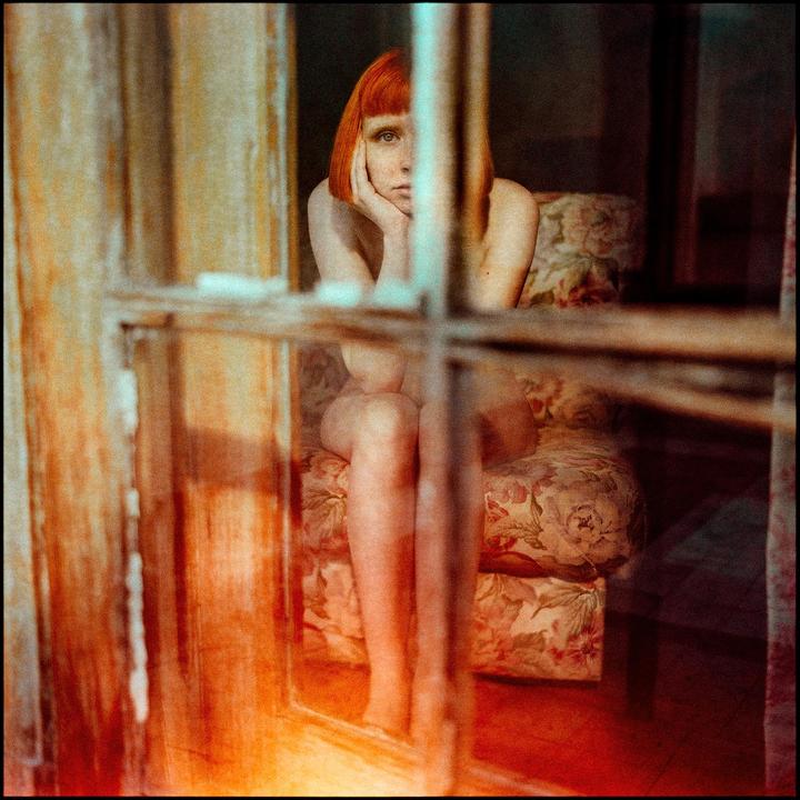 A woman with red hair sits on a floral armchair, seen through a window.