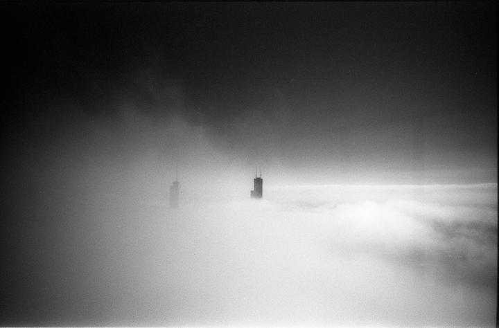 Two skyscrapers emerge from a thick layer of fog under a dark sky.
