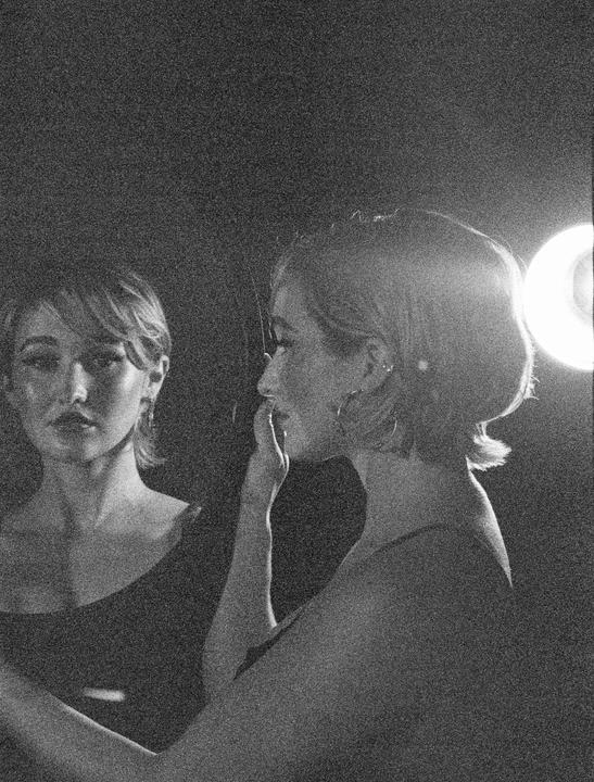 A woman is visible in profile with her reflection in a mirror, illuminated by a bright light source.
