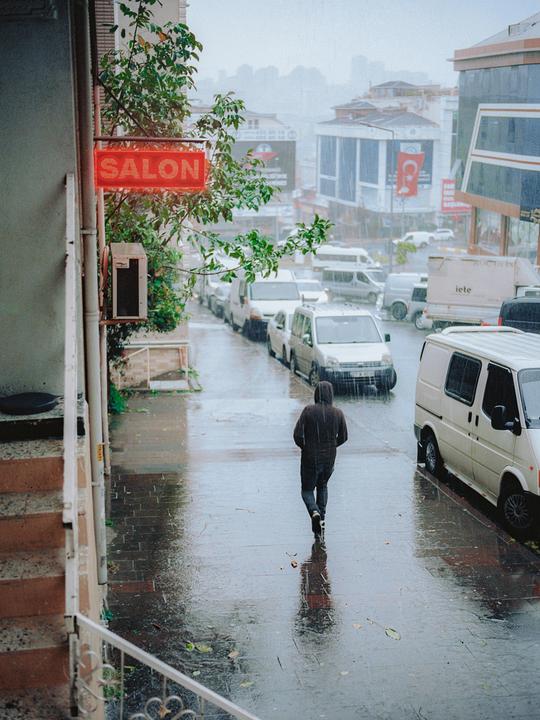 A person walks away from the camera on a wet street lined with parked vehicles and buildings, with a red "SALON" sign visible on the left.