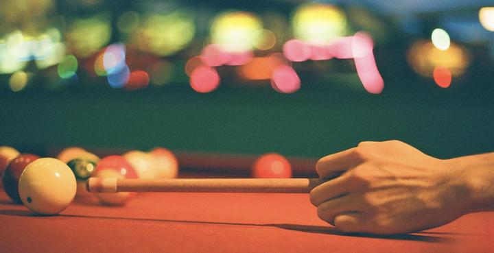 A hand holds a pool cue, aiming at a white cue ball on a red pool table with other balls scattered around.
