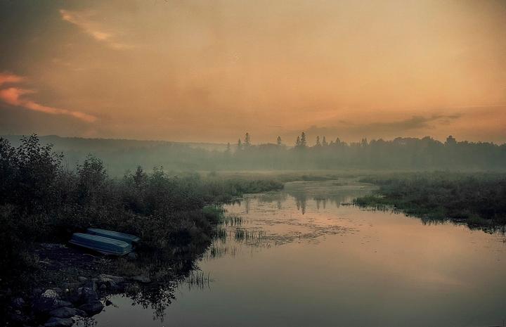 Two small boats rest on the bank of a misty river at sunset, with a forest in the background.