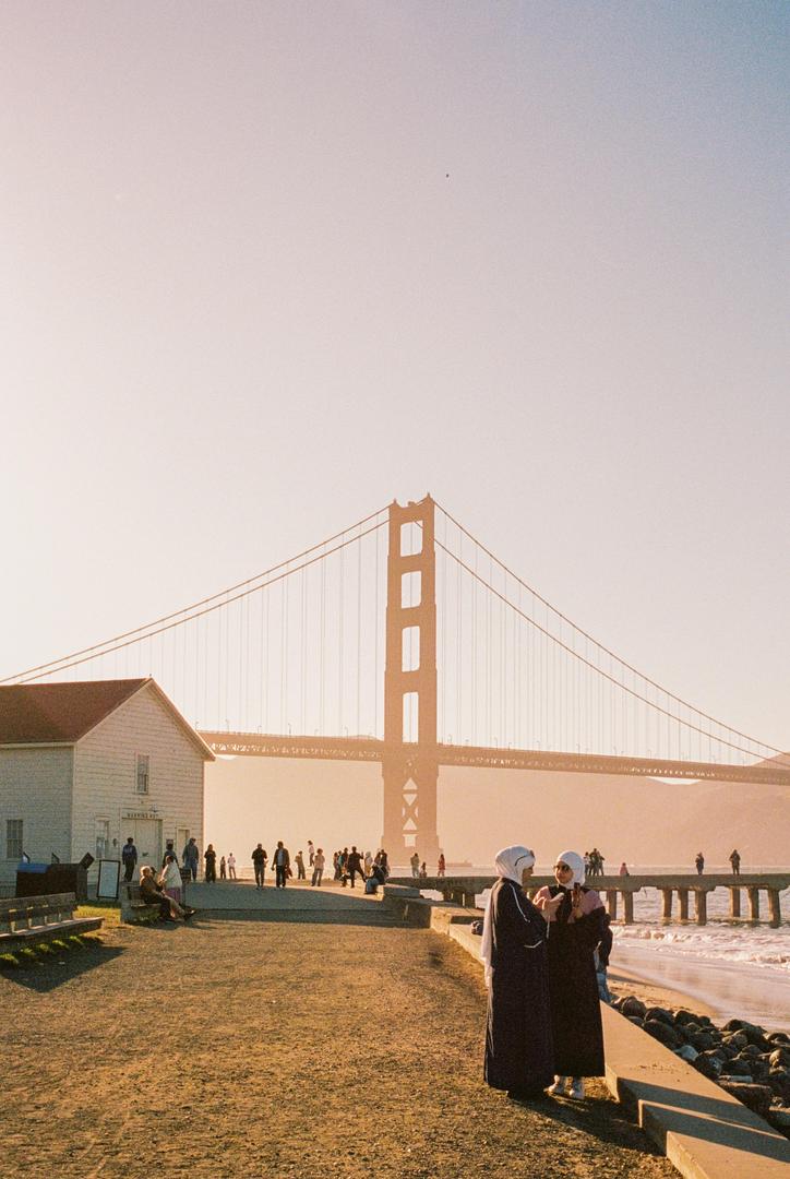 Two women stand on a beach with a bridge and a building in the background.