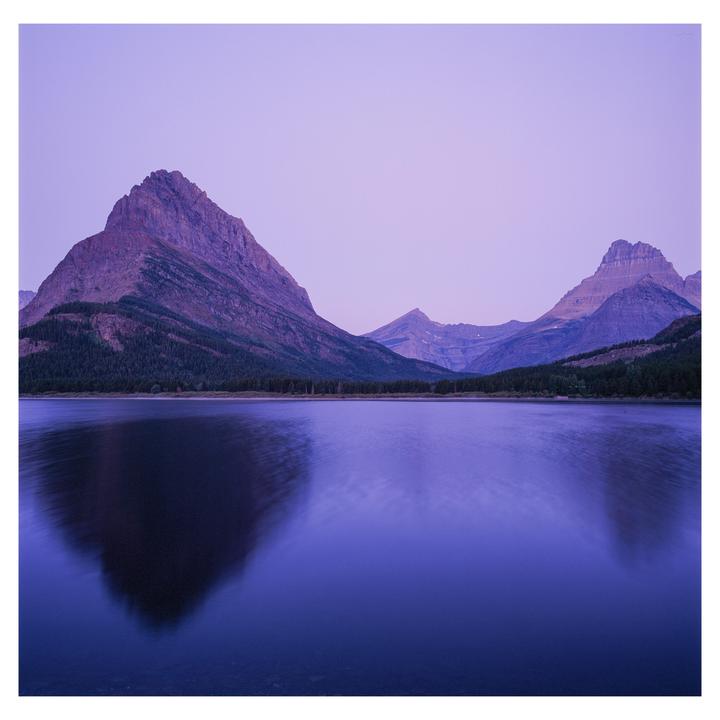 A calm lake reflects mountains and a forest under a purple sky.
