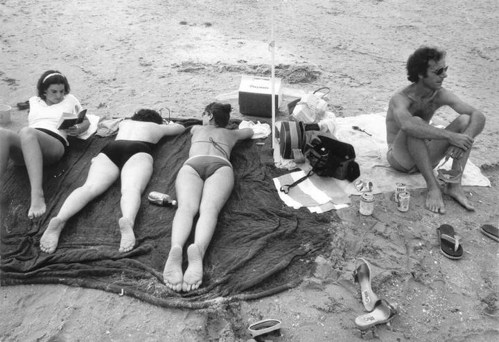 Three women and a man relax on a sandy beach with a cooler and drinks.