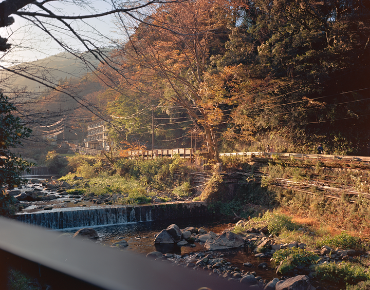 A river with small waterfalls flows past a road and buildings at the base of a mountain covered in autumn trees.