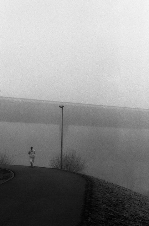 A person walks along a winding road next to a body of water on a foggy day.