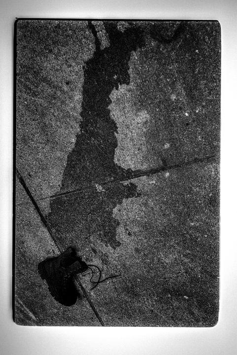 A single boot with an untied shoelace lies on a textured ground with a large dark stain.