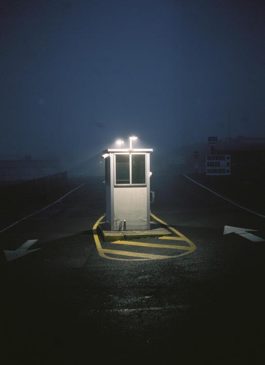 A small, illuminated security booth stands in the middle of a foggy road at night, with arrows painted on the asphalt.