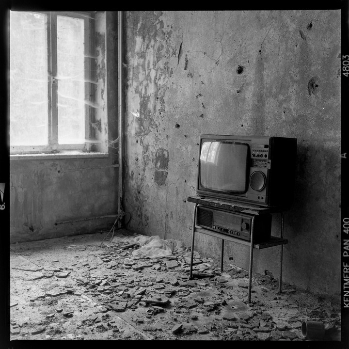 An old television set sits on a stand in a derelict room with a broken window and debris on the floor.