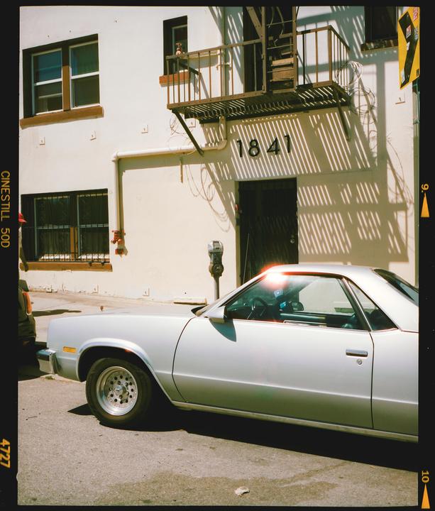 A silver car is parked on a street in front of a building with an external staircase and strong shadows.