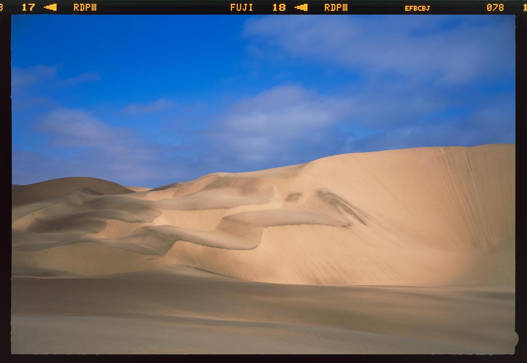 A vast desert landscape features rolling sand dunes under a partly cloudy blue sky.