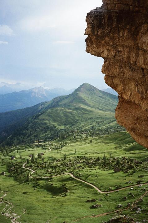 A winding path leads through a green valley with mountains in the background, framed by a rock formation on the right.