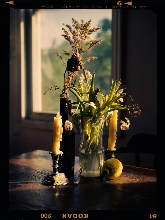 A still life composition features a vase of flowers, candles, and a lemon on a wooden table next to a window.