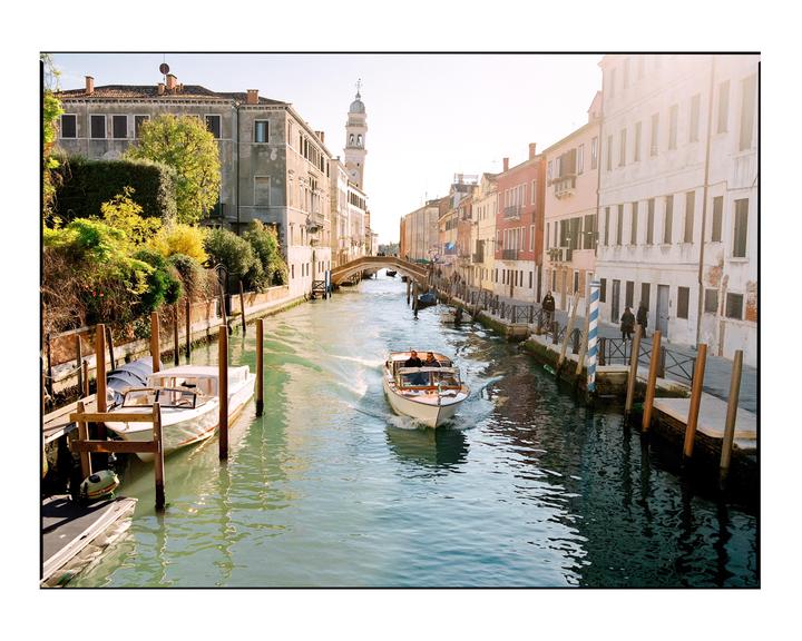 A boat with two people travels down a canal lined with buildings and other boats.