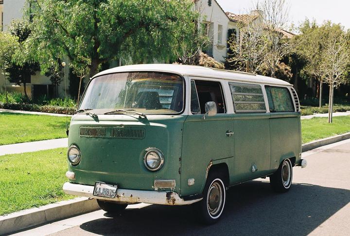 A green and white van is parked on a residential street next to a grassy area and trees.