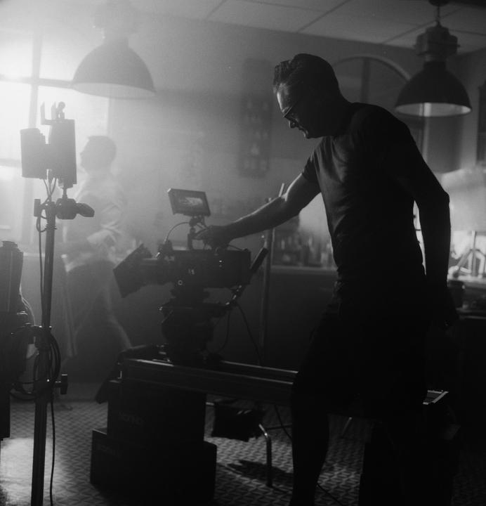 A man adjusts a camera on a dolly in a hazy room with another man in the background.