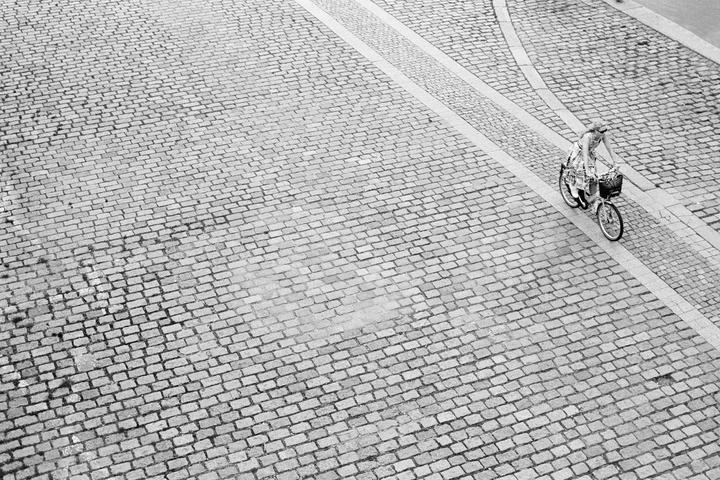 A woman rides a bicycle with a basket on a cobblestone street.